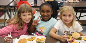 Three young girls smiling at the camera as they eat lunch in a school