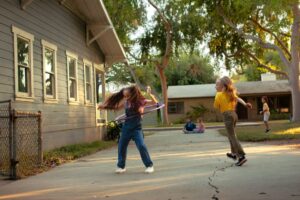 Two children playing in the yard of a home on a quiet, suburban street. The one with red hair is twirling a hula hoop.