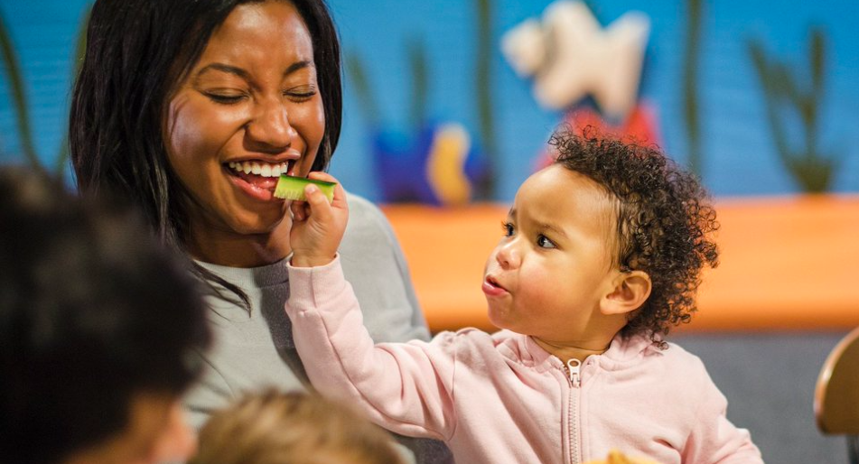 Young toddler feeding smiling mother at daycare center